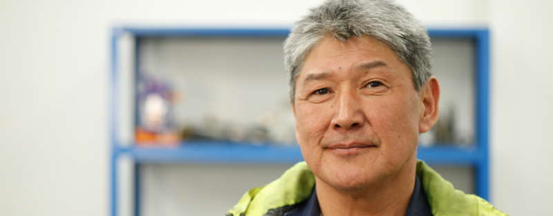 A person with gray hair stands in front of a blue shelf, wearing a yellow jacket.