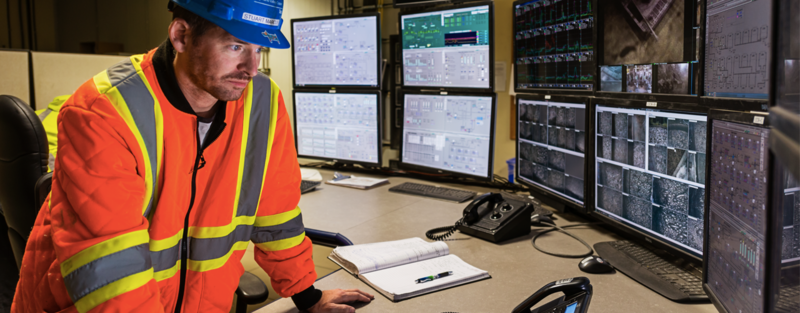 A worker in safety gear monitors multiple screens in a control room, with a notebook on the desk.