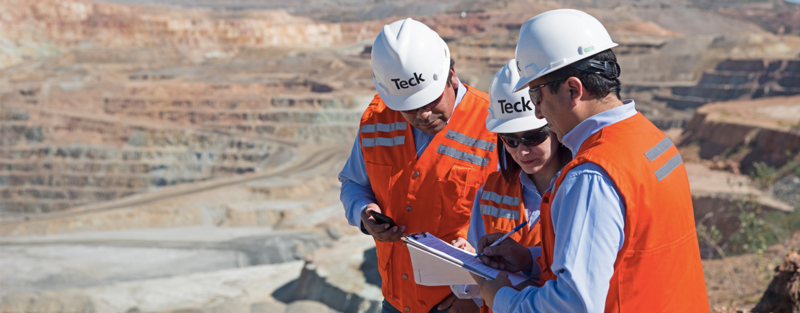 Three workers in safety gear and hard hats reviewing documents at a mining site.