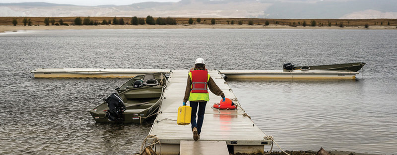 A person in safety gear and helmet carries a yellow container on a dock with boats on a lake.
