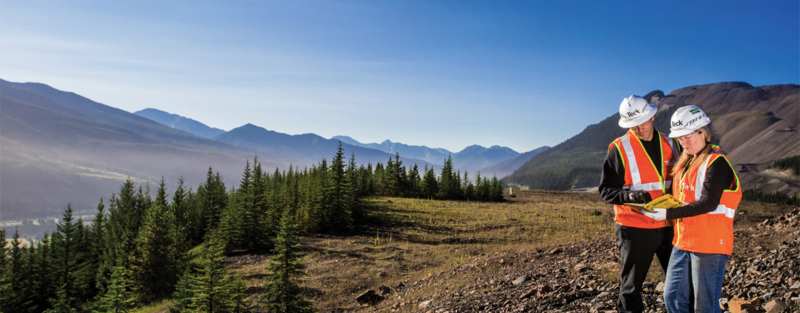 Two workers in safety gear examining plans on a mountainside with a forest and mountain range behind them.