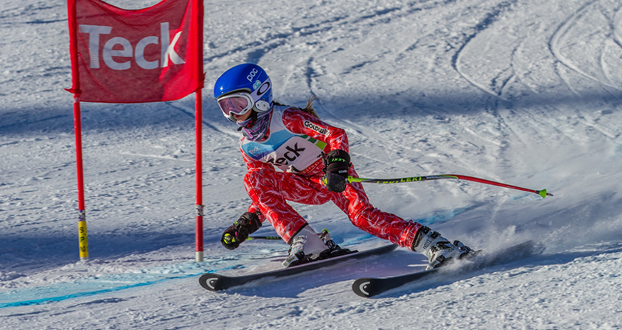 Young skier in red suit and blue helmet racing around a gate on a snowy slope.