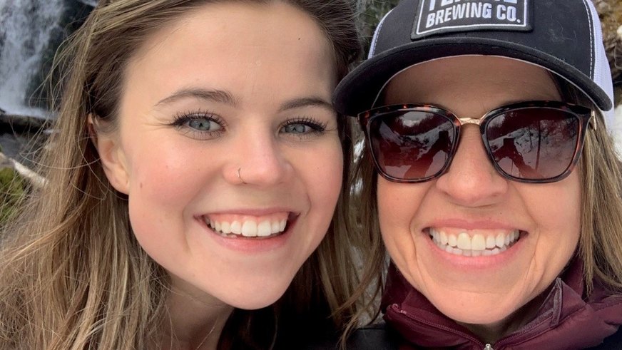 Two women smiling, one wearing sunglasses and a cap, with an outdoor background.