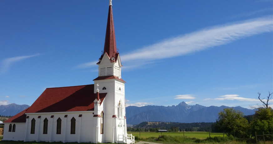 A white church with a red roof and steeple stands against a backdrop of mountains and blue sky.