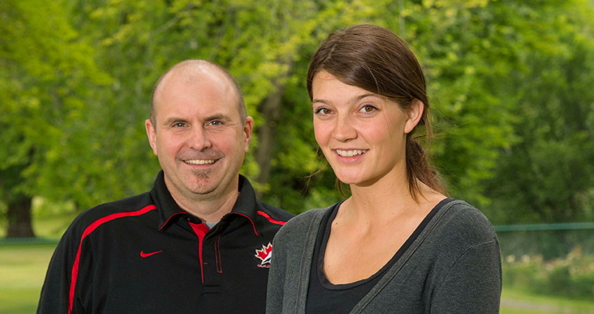 Man and woman smiling outdoors with trees in the background.