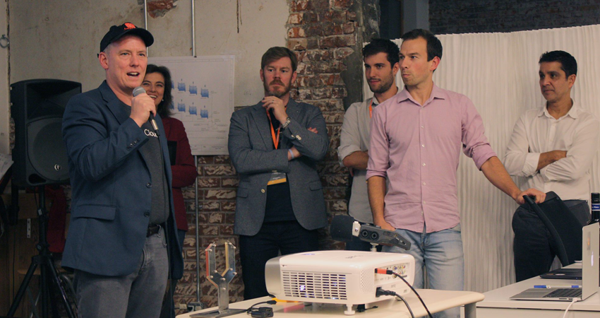 Man speaks into microphone, others look on, in a casual indoor setting with projector.