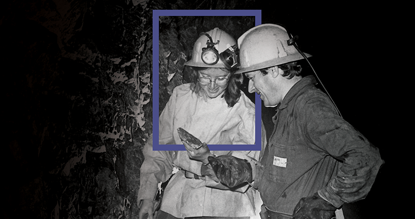 Two miners wearing helmets, examining a rock sample inside a dark cave or underground environment.