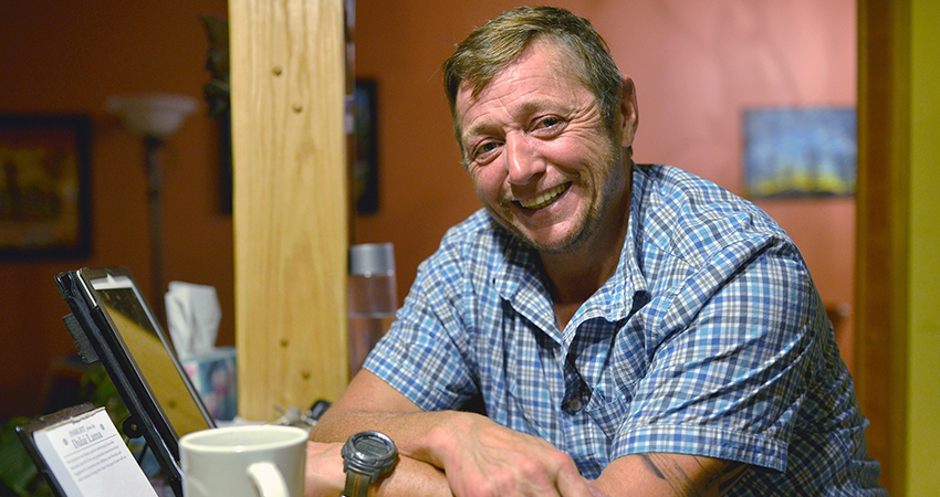 Man in plaid shirt sitting at a table, smiling, with a tablet and a coffee mug in front of him.