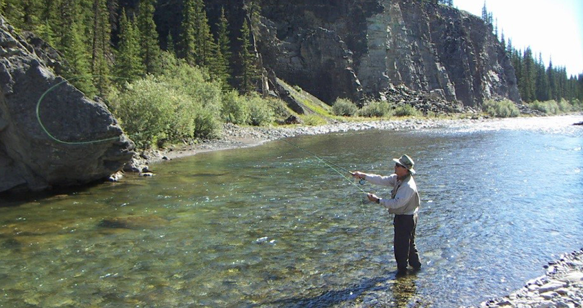 Person fly fishing in a clear river with rocky banks and forested hills in the background.
