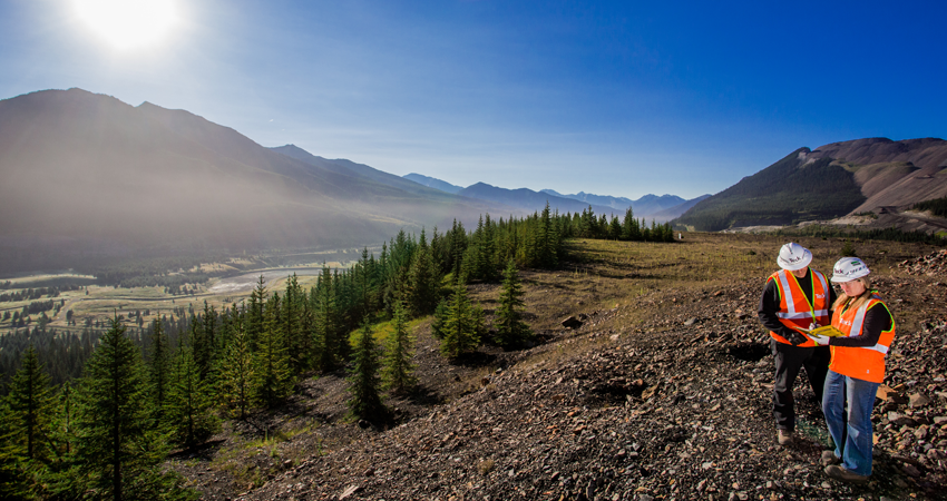 Two workers in safety vests and helmets overlook a mountainous landscape with forest and valley.