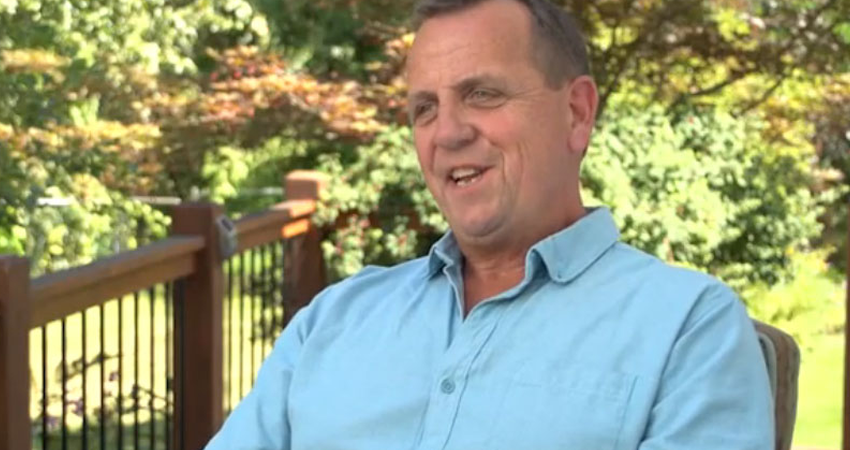 Man in a light blue shirt smiling outdoors with trees and a wooden fence in the background.