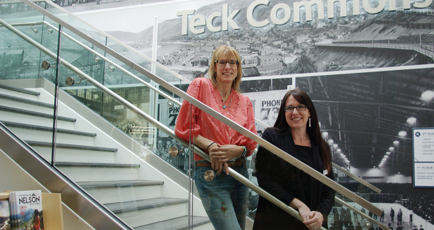 Two women smiling and posing on stairs in front of a mural with a 'Teck Commons' sign.