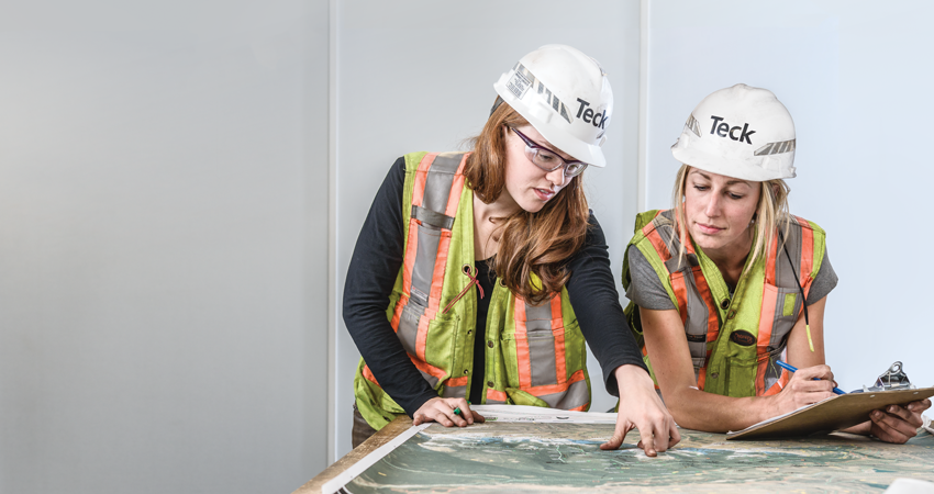 Two workers wearing hard hats and safety vests examine a detailed map on a large table.