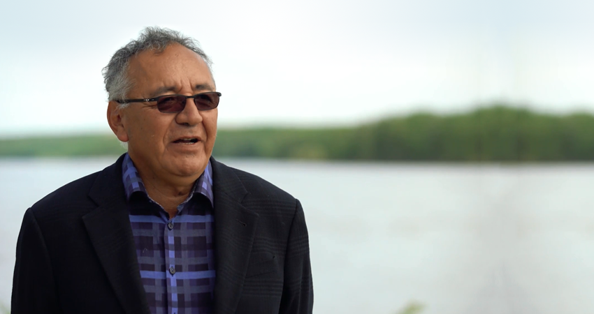 Man in glasses and a suit speaking with a lake and trees in the background.
