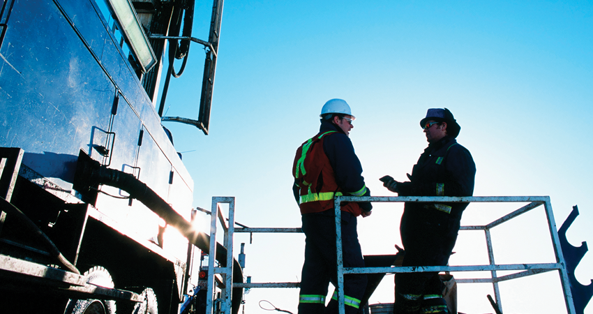 Two construction workers in safety gear talking next to a large vehicle against a clear blue sky.