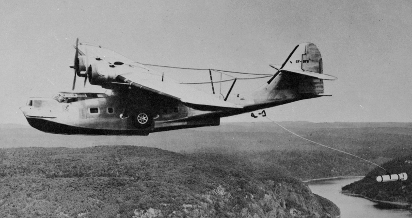 Vintage seaplane in flight over a river and forested landscape, towing an object.