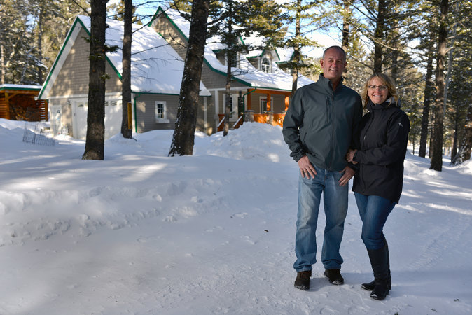 Couple standing in snow in front of a cozy cabin surrounded by trees in a winter landscape.