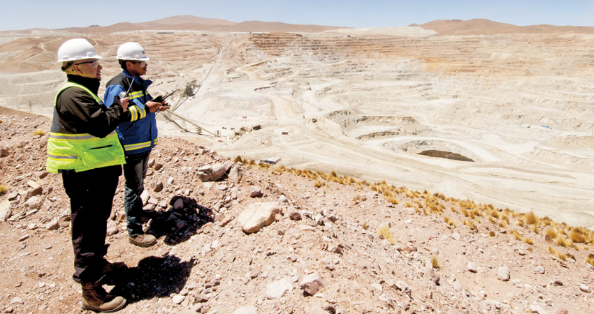 Two workers in safety gear overlook a large, open-pit mine with terraced levels and rocky terrain.
