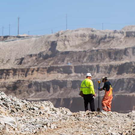 Two workers in safety gear stand with equipment at a rocky construction site.