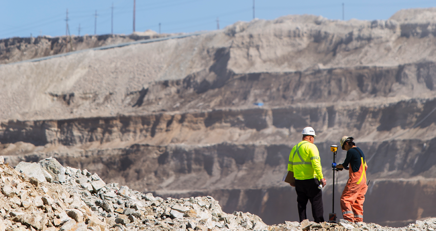 Two surveyors in safety gear working at a large quarry site with rocky terrain and steep hills.