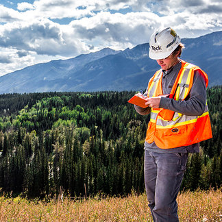 Worker in safety gear using a device in a mountainous forest area under a cloudy sky.