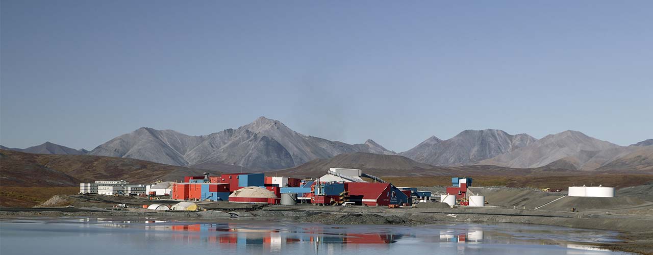 Mining facility with colorful buildings, mountains in the background, and a water body in the foreground.